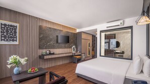 Habitación de hotel moderna con una cama grande, televisor de pantalla plana, una mesita con un florero y fruta, y una zona de estar con un banco.