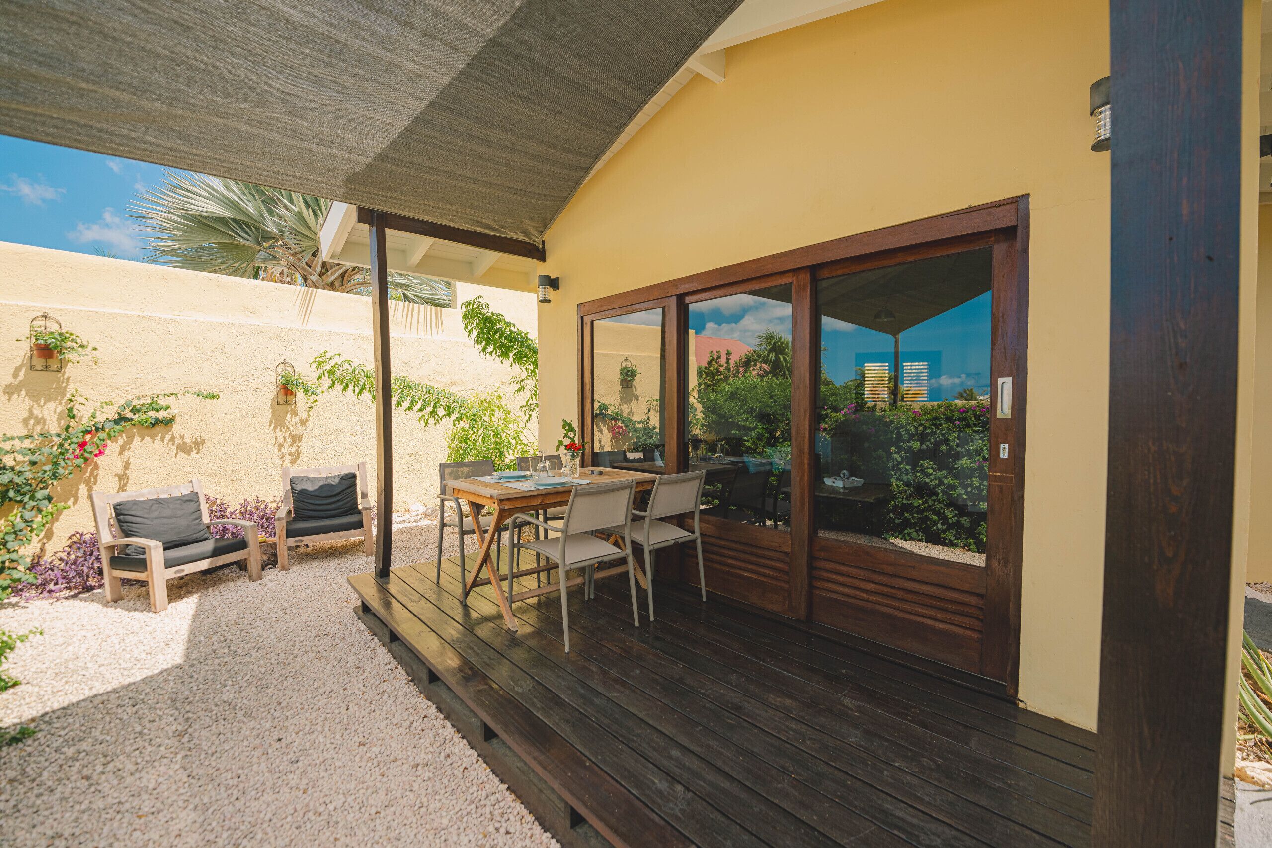 A wooden deck with a table and chairs, a covered patio area, and a sliding glass door leading to a garden.