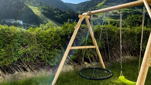 Eine Holz-Schaukel mit grüner Sitzfläche vor einer Kulisse aus saftig grünen Hügeln und blauem Himmel.
