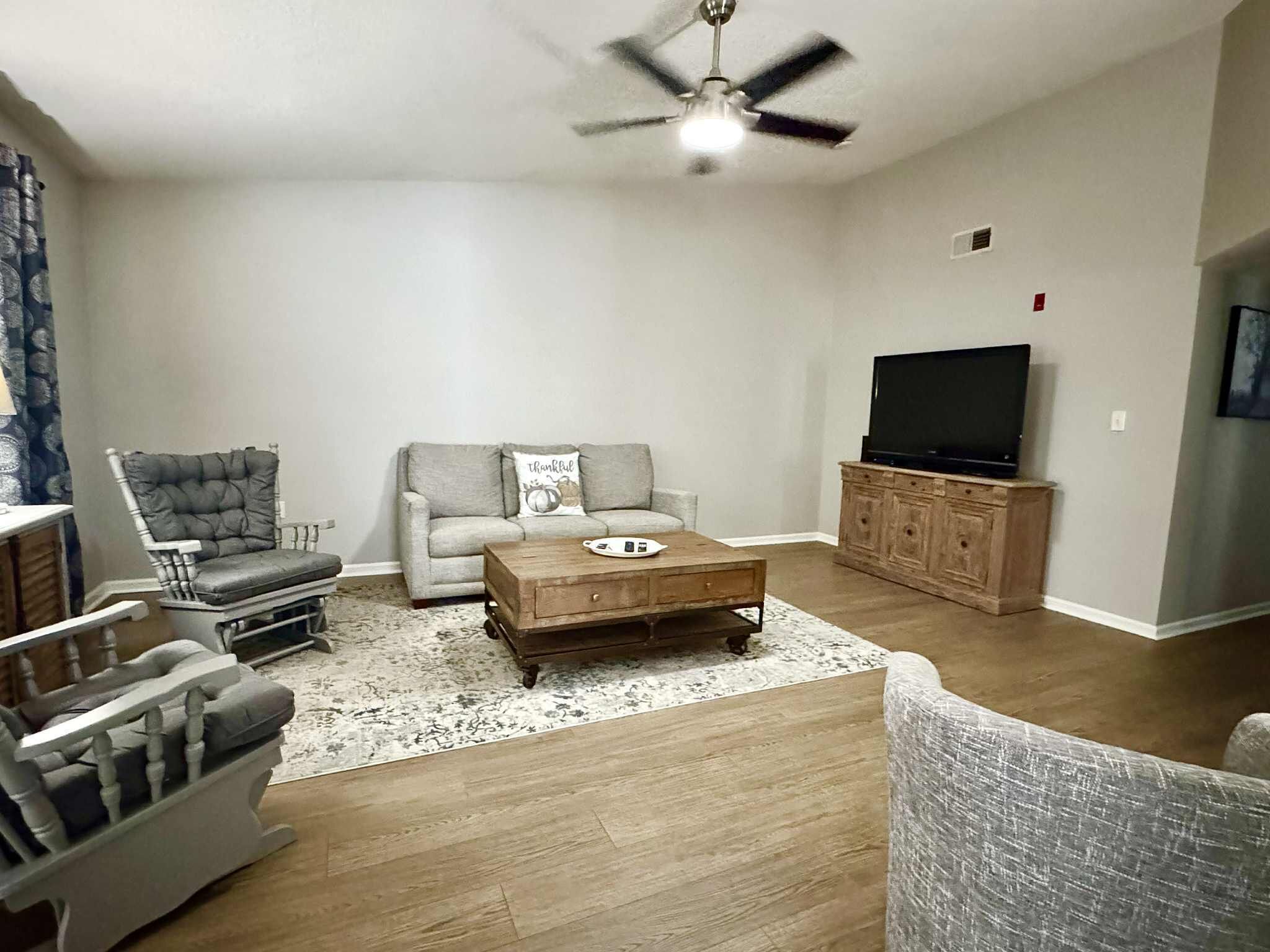 Ein Wohnzimmer mit grauem Sofa, einem Holztisch, einem grauen Sessel, einem Fernseher auf einer Holzkommode und einem Deckenventilator.