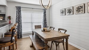 A dining area with a wooden table, benches, and chairs. There are curtains, a hanging light fixture, and framed artwork on the wall.