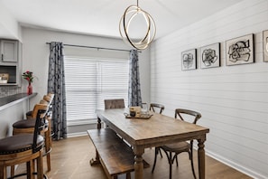 Un comedor con mesa de madera, bancos y sillas. Hay cortinas, una lámpara colgante y cuadros en la pared.
