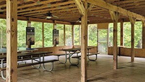 Un área de picnic techada con bancos y mesas de madera, rodeada de amplias ventanas que ofrecen vistas a una zona boscosa.