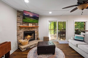 Una sala de estar con chimenea de piedra, un televisor montado encima y un sofá, con un área de comedor visible a través de puertas de cristal.