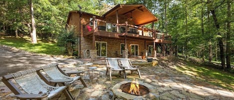 A cabin with a stone fire pit, outdoor seating, and a deck overlooking a forest.