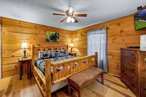 Une chambre aux murs lambrissés, comprenant un lit, deux lampes de chevet, un ventilateur de plafond, un banc en bois, une commode et un téléviseur fixé au mur.