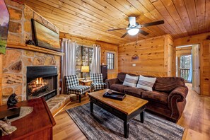 L’intérieur d’un chalet accueillant, avec une cheminée en pierre, des murs en bois, un canapé en cuir marron et un ventilateur de plafond.