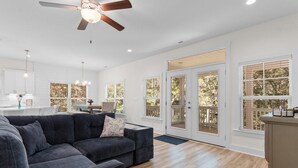 Ein geräumiges Wohnzimmer mit einem dunkelgrauen Ecksofa, einem Deckenventilator und großen Fenstern mit Blick ins Freie.