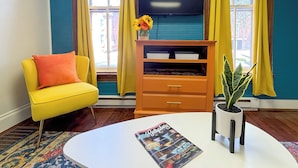 Ein Zimmer mit einem gelben Sessel, einem weißen Couchtisch mit einer Zeitschrift, einem Holzschrank und einer Topfpflanze.