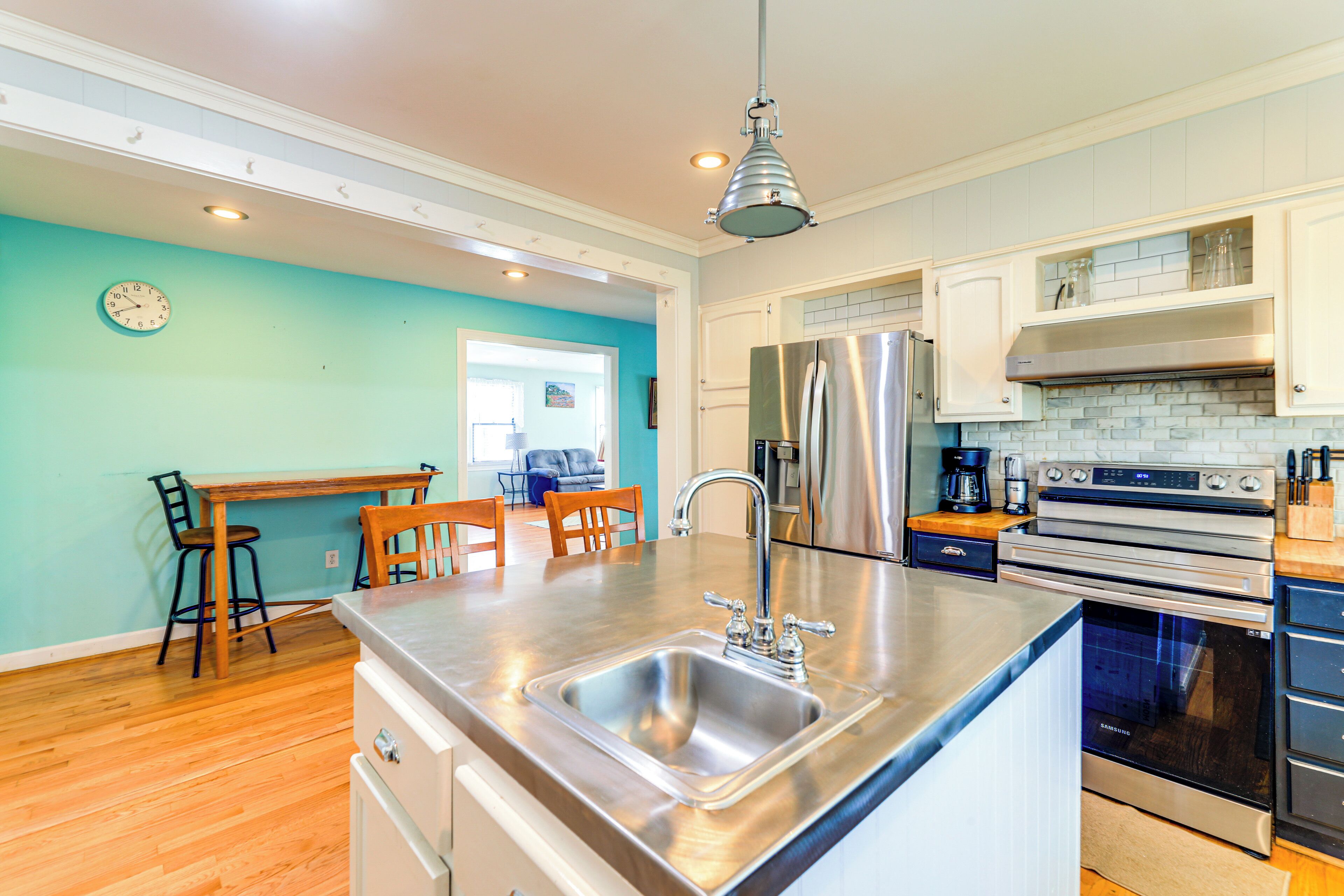 A modern kitchen with stainless steel appliances, a large island with a sink, wooden chairs, and a teal wall.