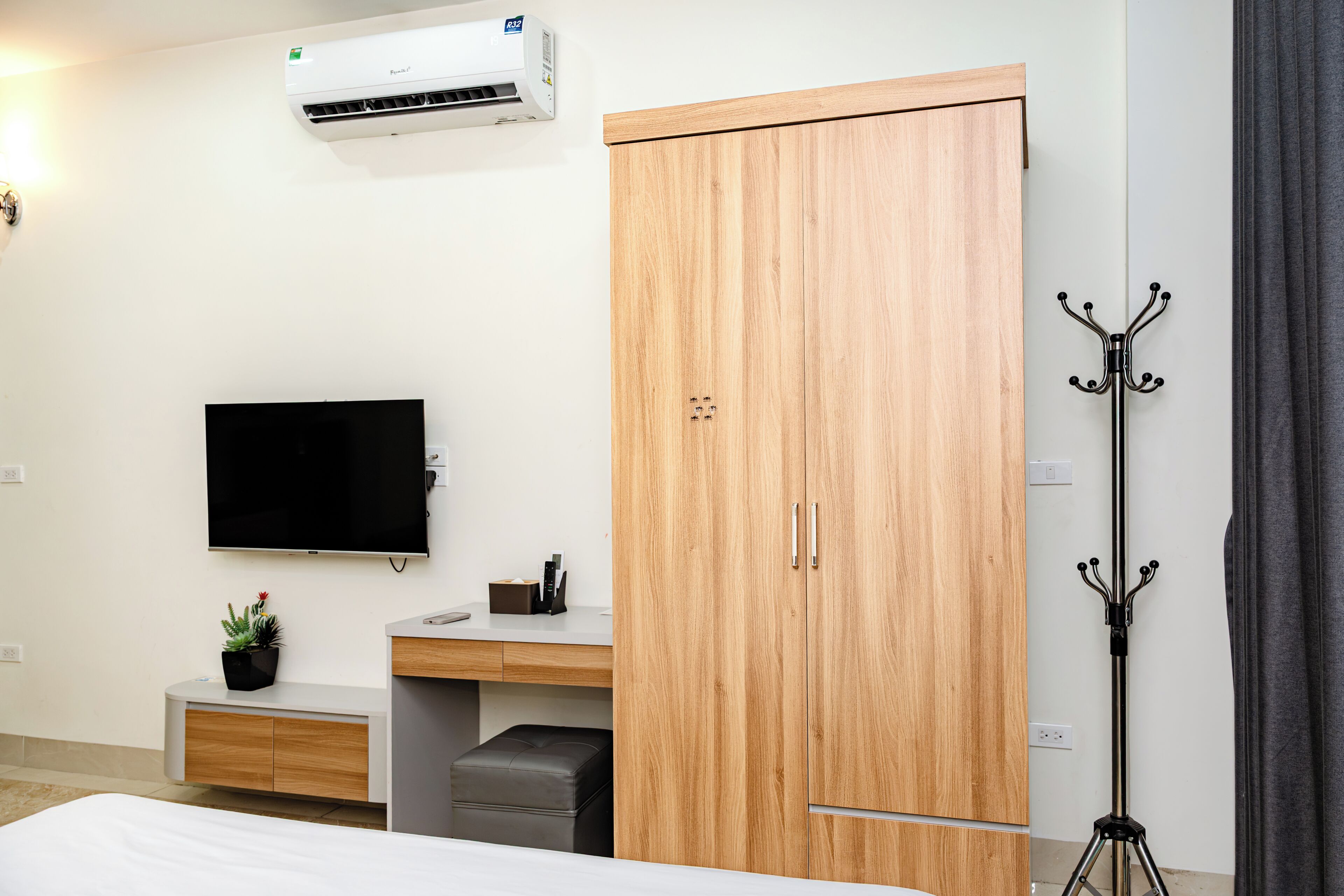 A hotel room with a bed, a desk with a flat-screen TV, a wardrobe, a coat rack, and a potted plant.