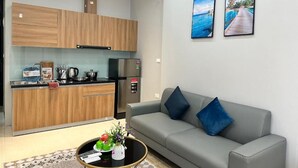 A modern kitchen with wooden cabinets, a black refrigerator, and a coffee machine. There is a grey sofa with blue cushions and a small round table with a vase and a book. Two framed pictures are on the wall. - Hoang Long Apartment (Hanoi)