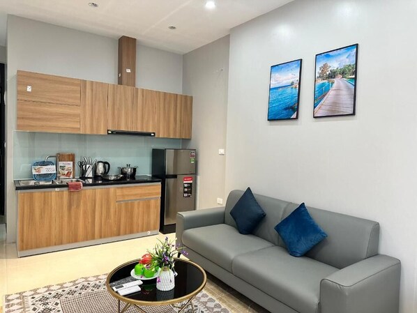 A modern kitchen with wooden cabinets, a black refrigerator, and a coffee machine. There is a grey sofa with blue cushions and a small round table with a vase and a book. Two framed pictures are on the wall. - Hoang Long Apartment (Hanoi)
