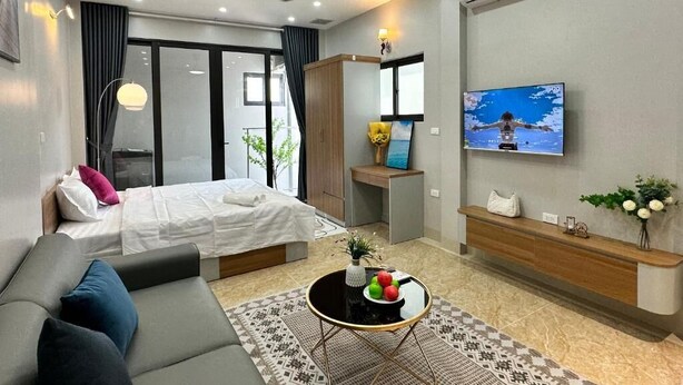 A modern hotel room with a bed, a sofa, a coffee table with fruit, a TV, and a ceiling fan. - Hoang Long Apartment (Hanoi)