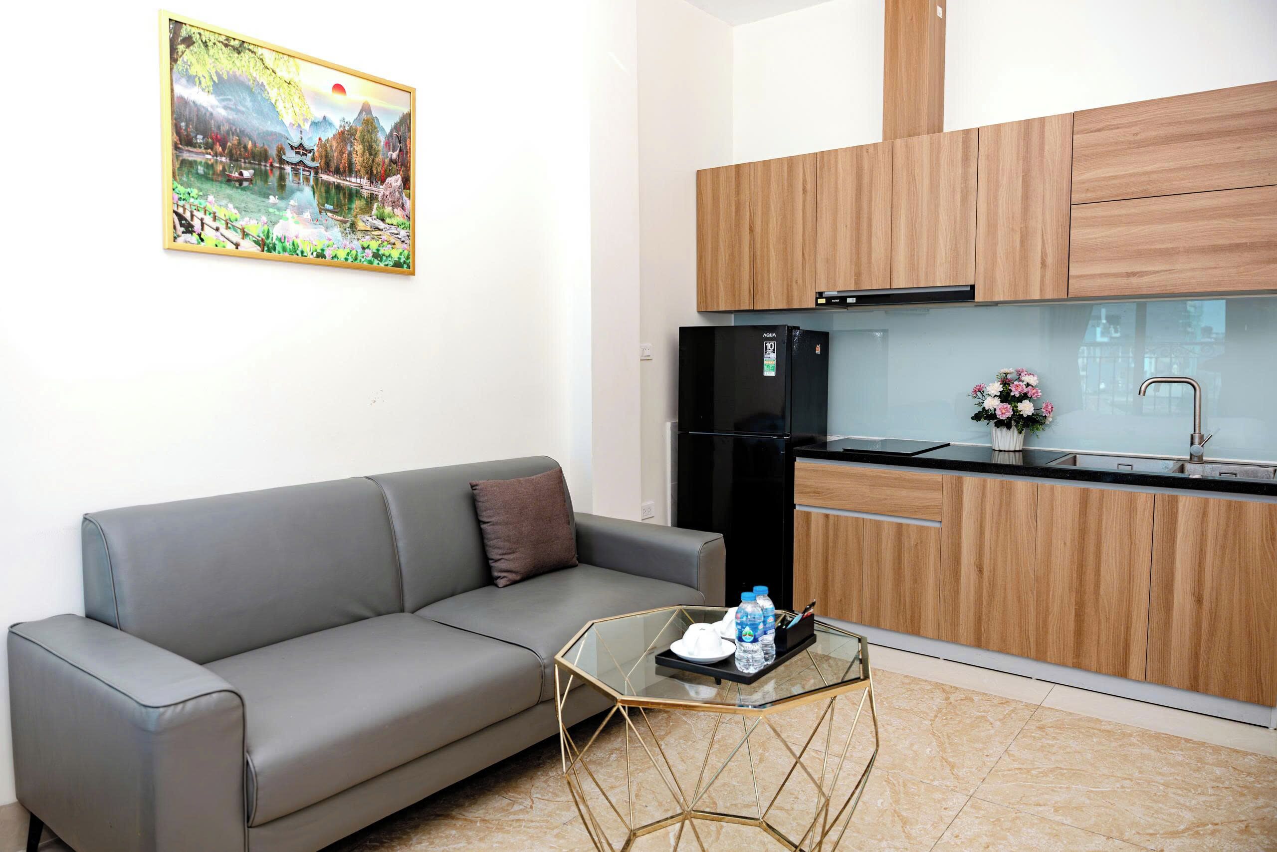 A modern living space with a grey sofa, a glass coffee table, wooden cabinets, and a kitchen area with a black refrigerator.