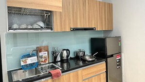 A compact kitchen with wooden cabinets, a stainless steel sink, a black refrigerator, and a dish rack. - Hoang Long Apartment (Hanoi)