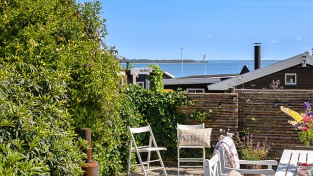 Una terraza de madera con sillas y una mesa blancas, rodeada de exuberante follaje y vista al mar.