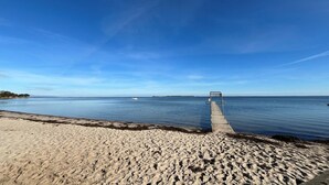 Una playa con un muelle de madera que se adentra en aguas tranquilas, un cielo despejado y tierra a lo lejos.