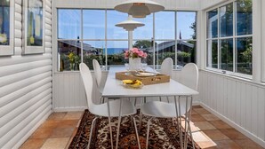 Un comedor con una mesa blanca, cuatro sillas, una lámpara colgante y vistas al exterior a través de amplios ventanales.