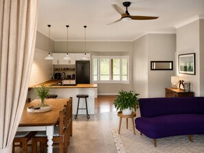 Ein modernes Wohnzimmer mit einem Holzesstisch, einem violetten Sofa und einer Küchenzeile mit weißen Schränken und einem schwarzen Kühlschrank.