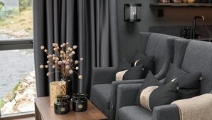 A modern living room with a grey sofa, a wooden side table with candles, and a view of a rocky coastline through large windows. - Hideaway Herdla 1 – unique and luxurious cabin 15 m above the sea near Bergen (Herdla)
