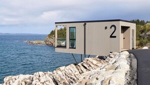 Une cabane moderne et minimaliste, dotée d’une grande fenêtre, située au bord d’une côte rocheuse et offrant une vue sur la mer.
