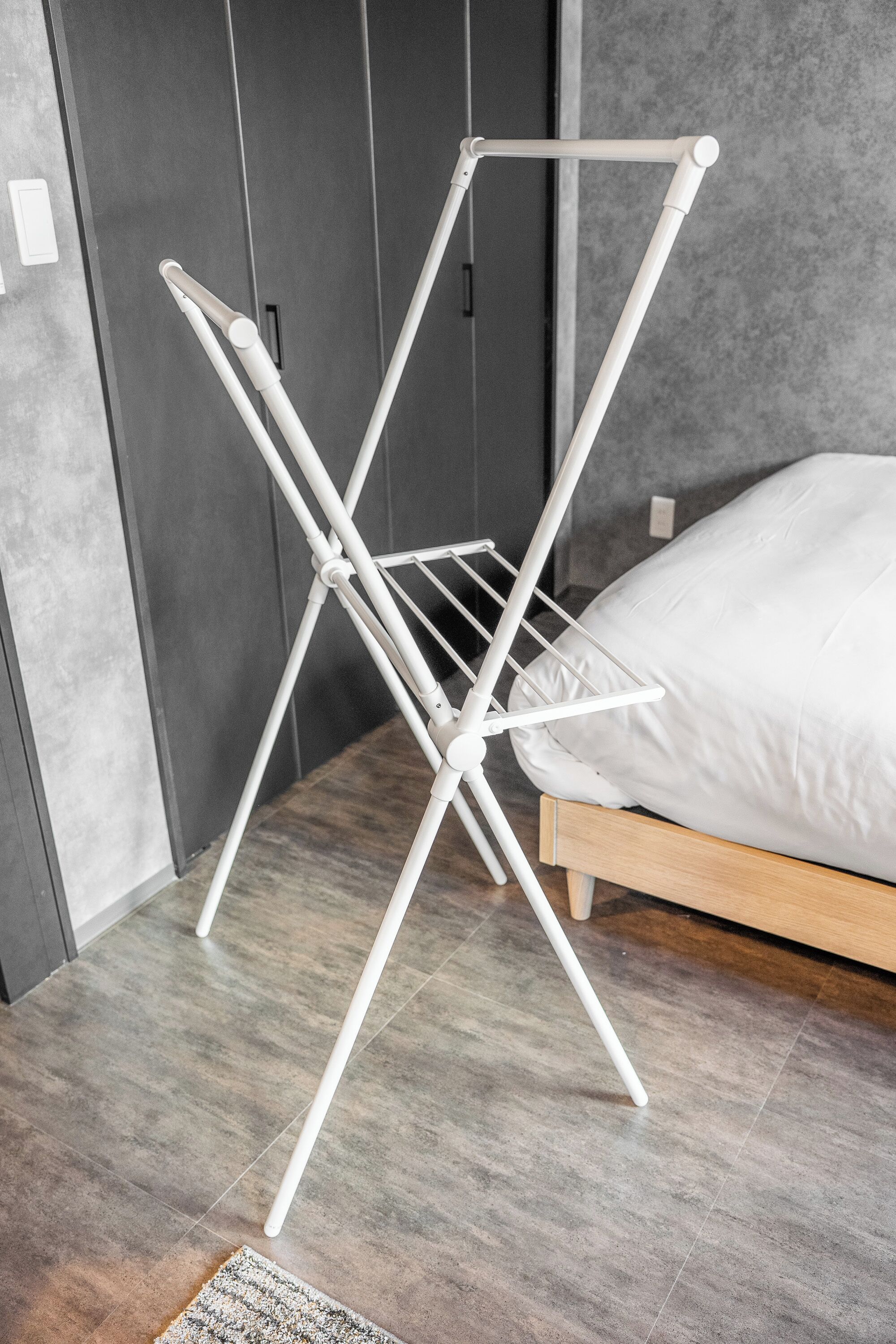 A white drying rack in a bedroom with a wooden bed and grey walls.