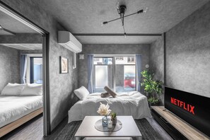 Une chambre d’hôtel moderne avec un lit, un téléviseur affichant « NETFLIX », une petite table avec un vase et une plante, et une fenêtre avec des rideaux.