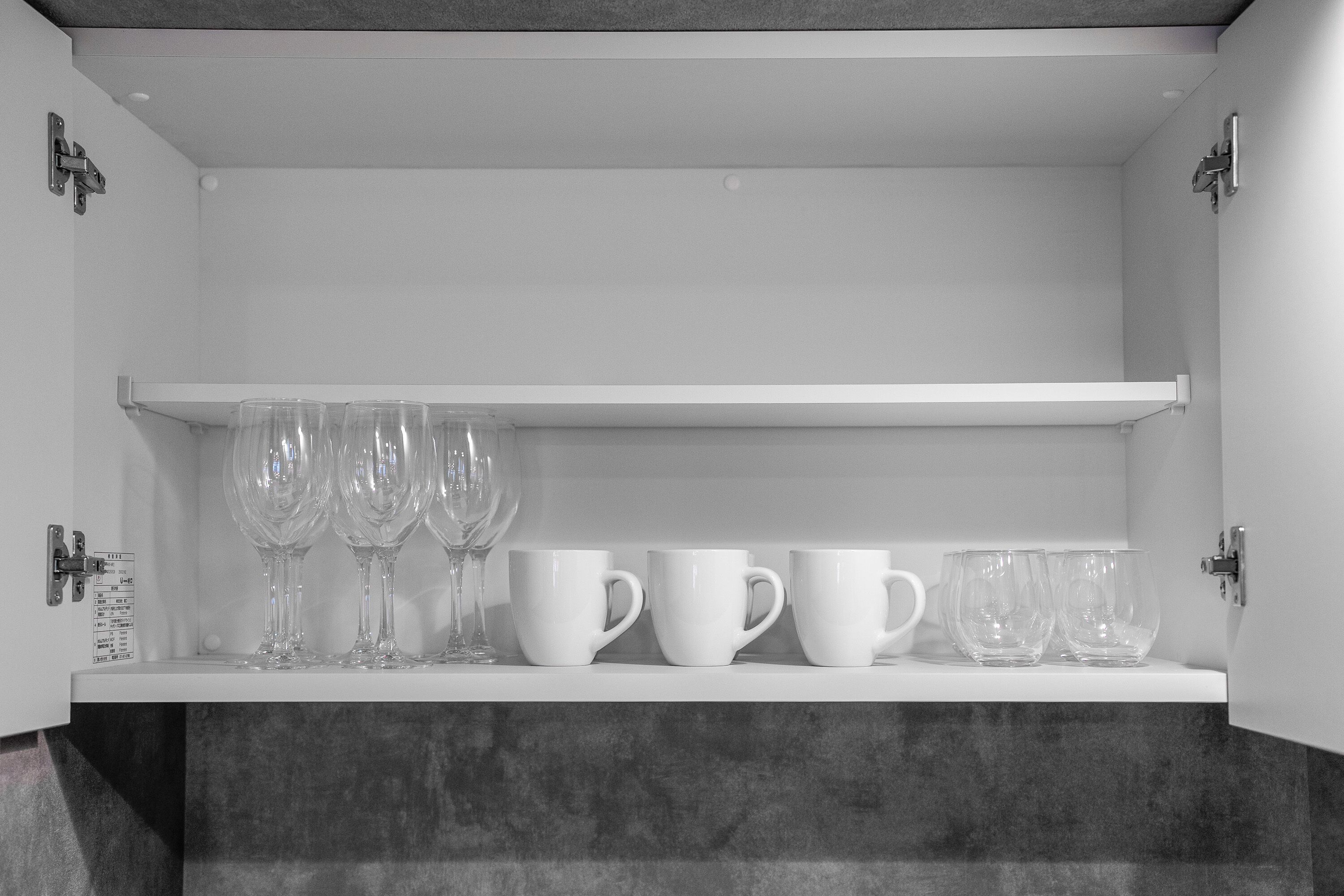 A white cabinet with shelves holding clear wine glasses and white mugs.