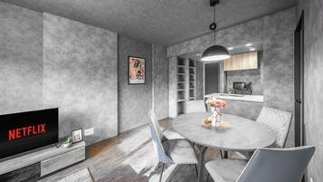 A modern living room with a flat-screen TV, grey upholstered chairs, a round dining table, and a kitchen area visible through an open doorway.