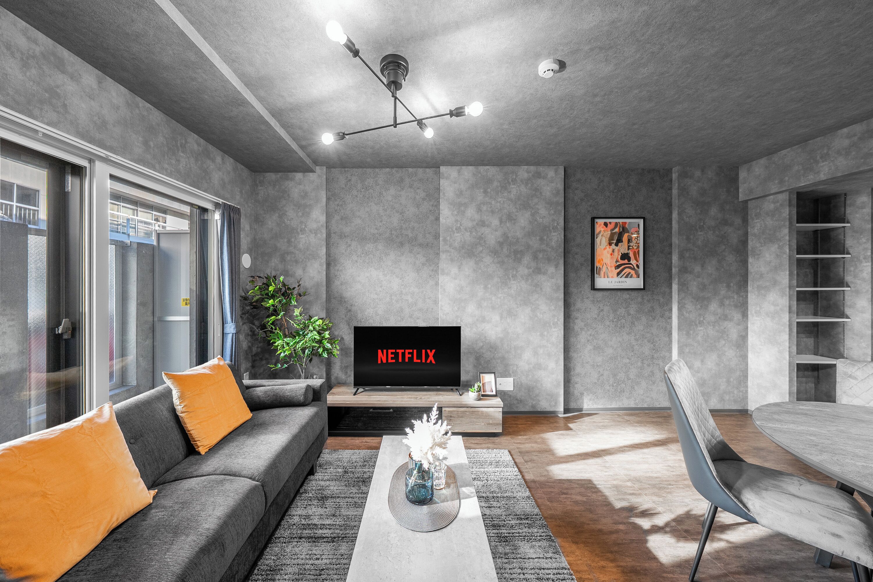 Moderni olohuone, jossa on harmaa sohva, maljakkoon asetettu kahvipöytä, seinälle ripustettu televisio, jossa näkyy teksti »NETFLIX», sekä ruokailutila, jossa on puinen pöytä ja tuolit.
