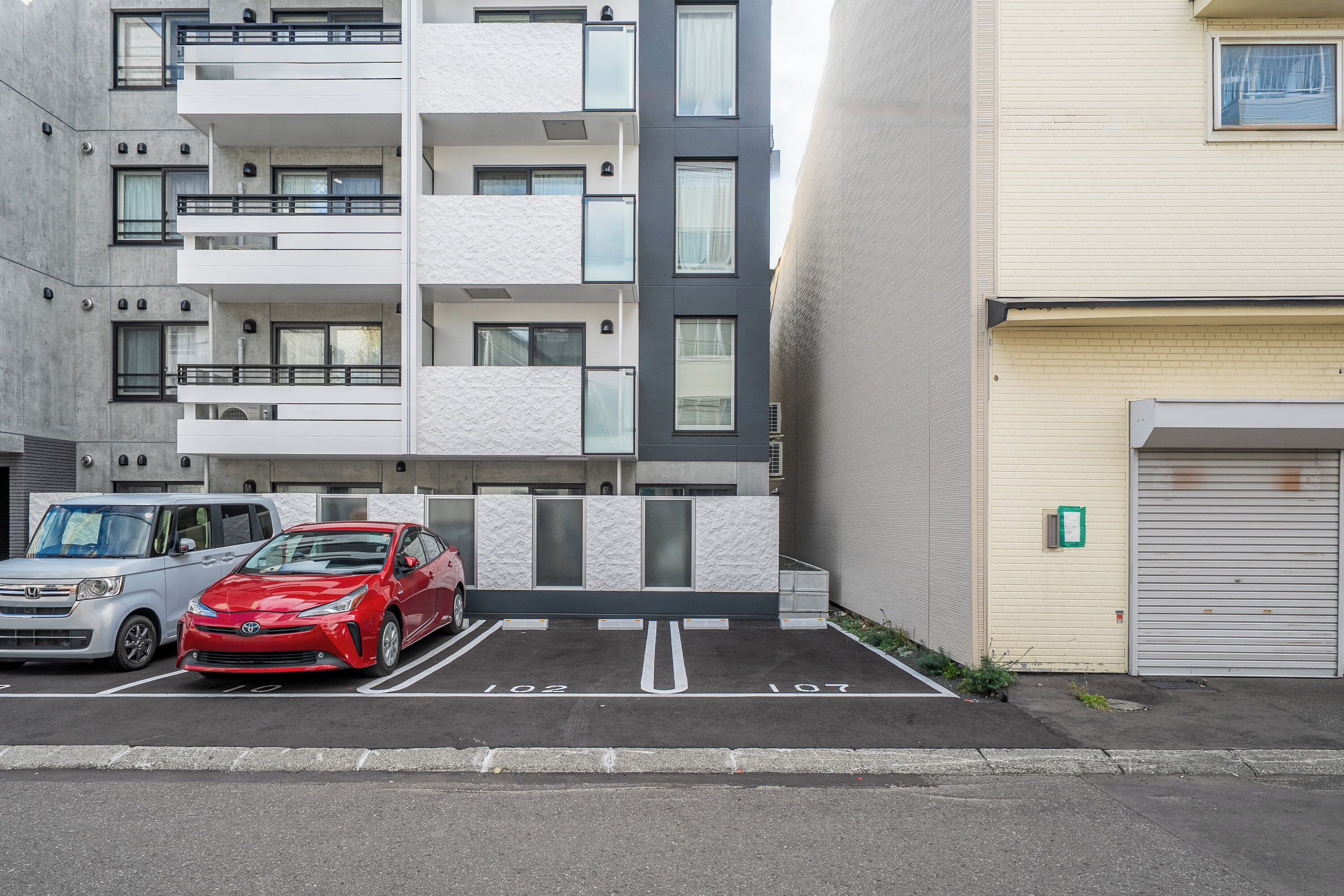 A parking lot with two cars, a red and a silver one, in front of a modern building with balconies and a garage door.