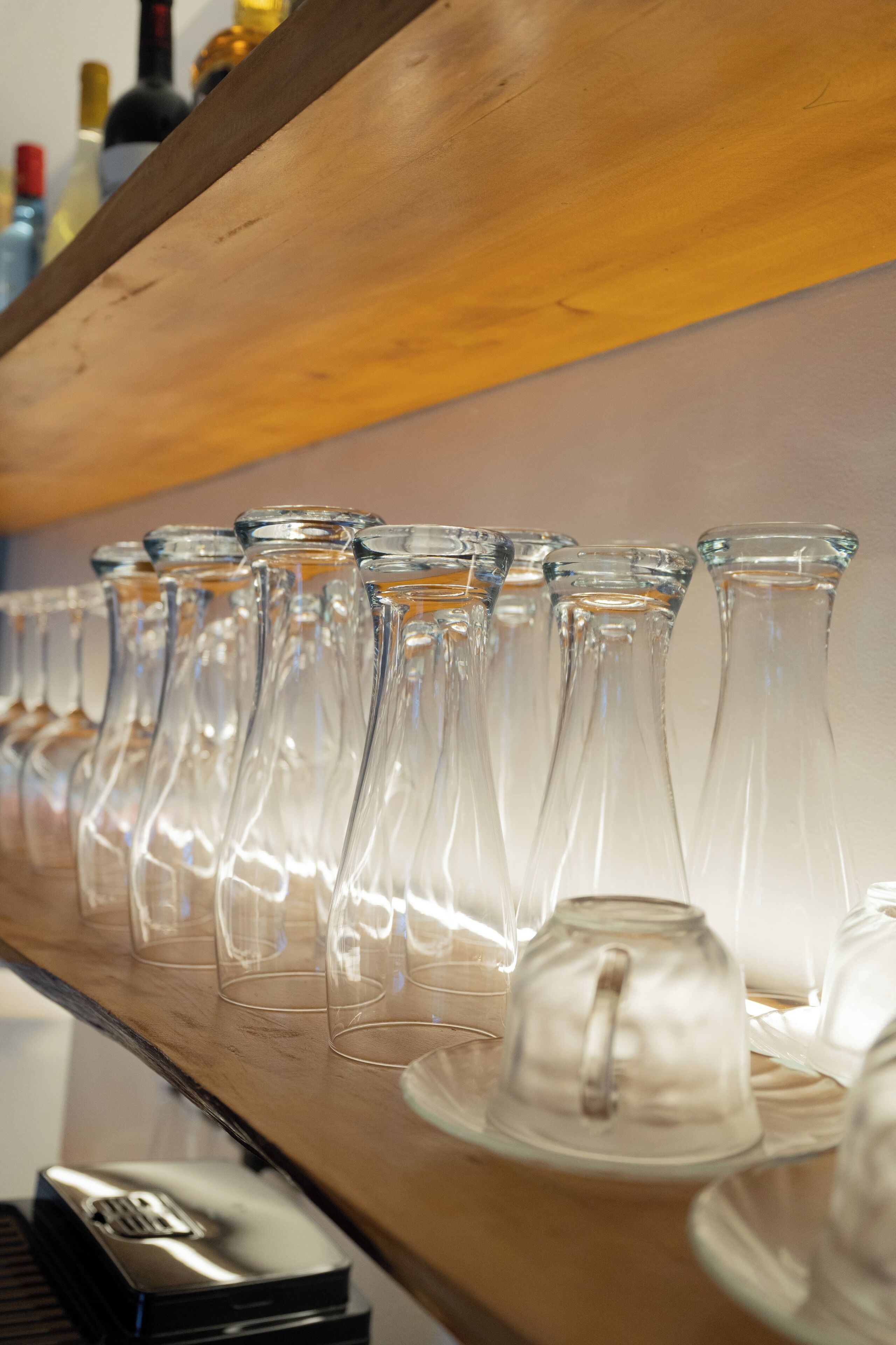 A bar area with a wooden shelf holding multiple clear glass wine glasses and a few white teacups.