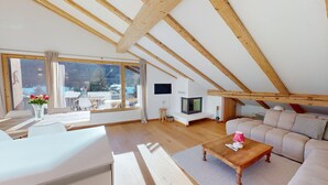 A spacious loft with a high ceiling, wooden beams, a large window, a television, a sofa, and a coffee table. - Charming holiday home in an excellent, quiet location in sun-drenched Pontresina (Pontresina)