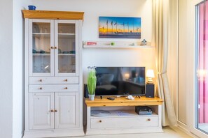 Une pièce avec un téléviseur posé sur un meuble en bois, une armoire aux portes en verre, un tableau accroché au mur et une fenêtre avec des rideaux.