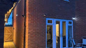 A brick building with a wooden gate, a sliding glass door, and a small patio area with a chair and table. - Comfortable 5 - Guest Abode - In Quiet Village (Grantham)