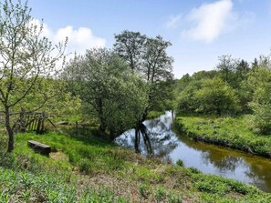 A serene riverside landscape with lush greenery and a wooden bench. - 5 Star Holiday Home in Grindsted (Grindsted)