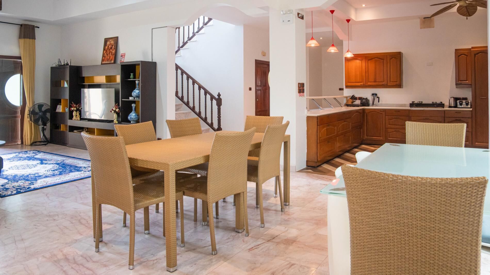 A modern kitchen with wooden cabinets, a dining area with wicker chairs, and a staircase leading to another level.
