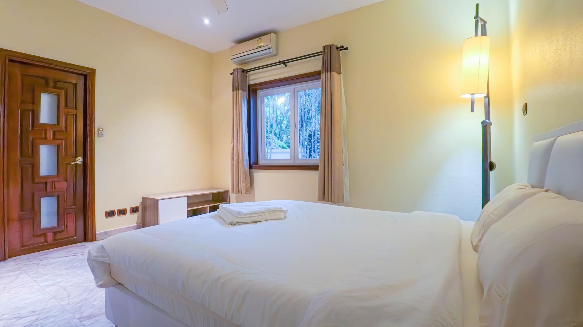 A hotel room with a bed, a window with curtains, an air conditioning unit, a bedside lamp, and a wooden door.