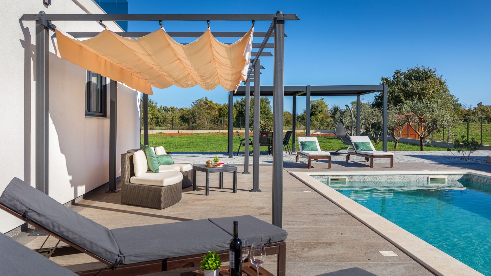 A poolside area with lounge chairs, a table with a bottle and glass, and a pergola with a canopy.