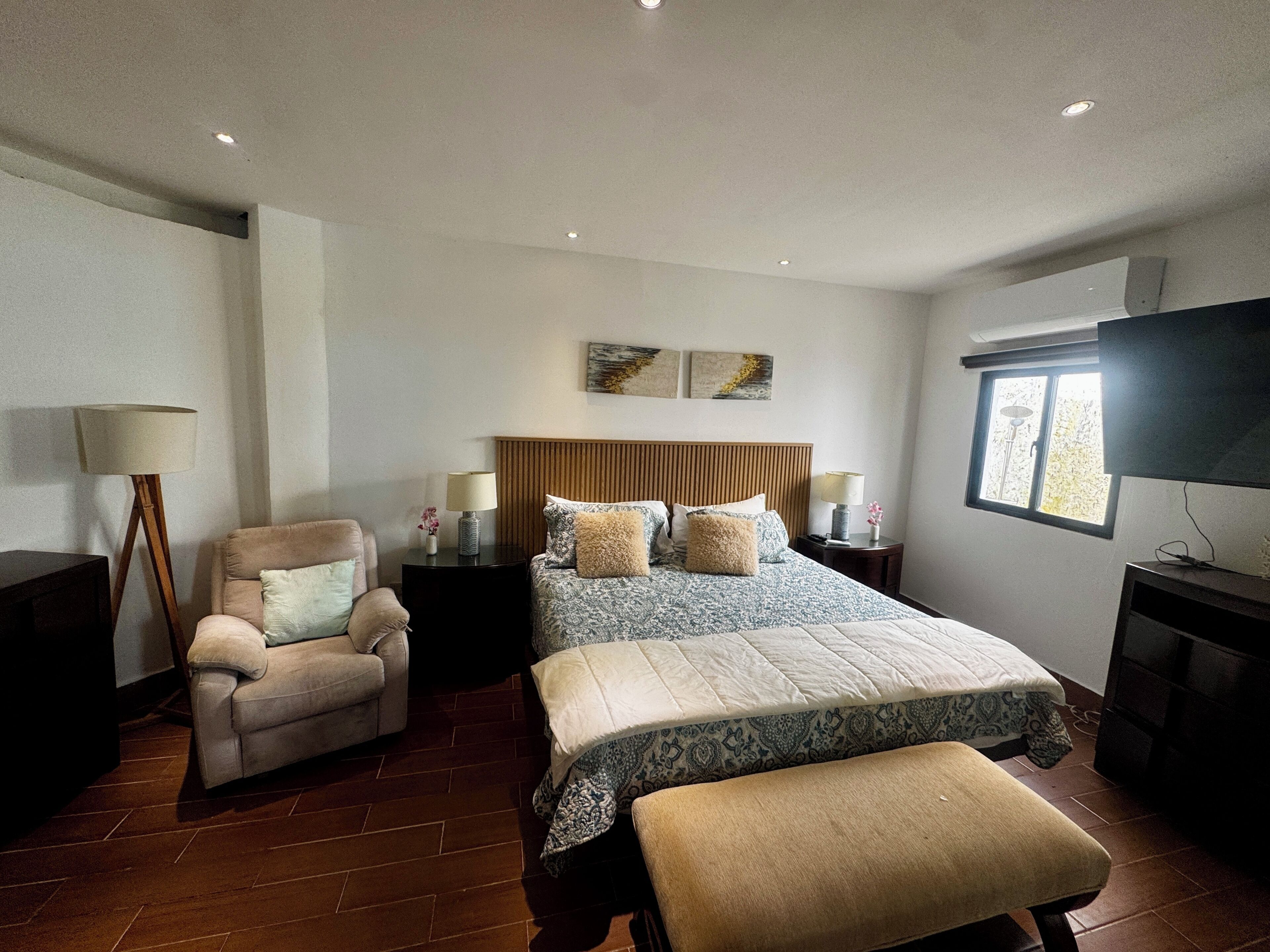A bedroom with a bed, a chair, a bedside table, a lamp, a TV, and a window.