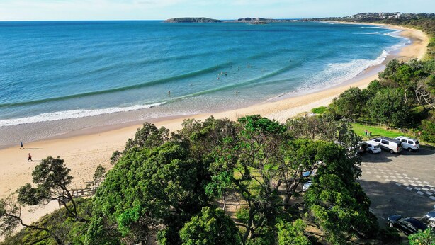 Une plage avec une eau d’un bleu limpide, un rivage sablonneux et un espace de stationnement où sont garées des voitures.