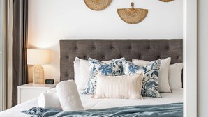 Un letto rifatto con cura, con una testiera grigia, biancheria da letto bianca e una coperta azzurra. Sopra il letto, tre arazzi tessuti, e una lampada da comodino è visibile sul comodino.