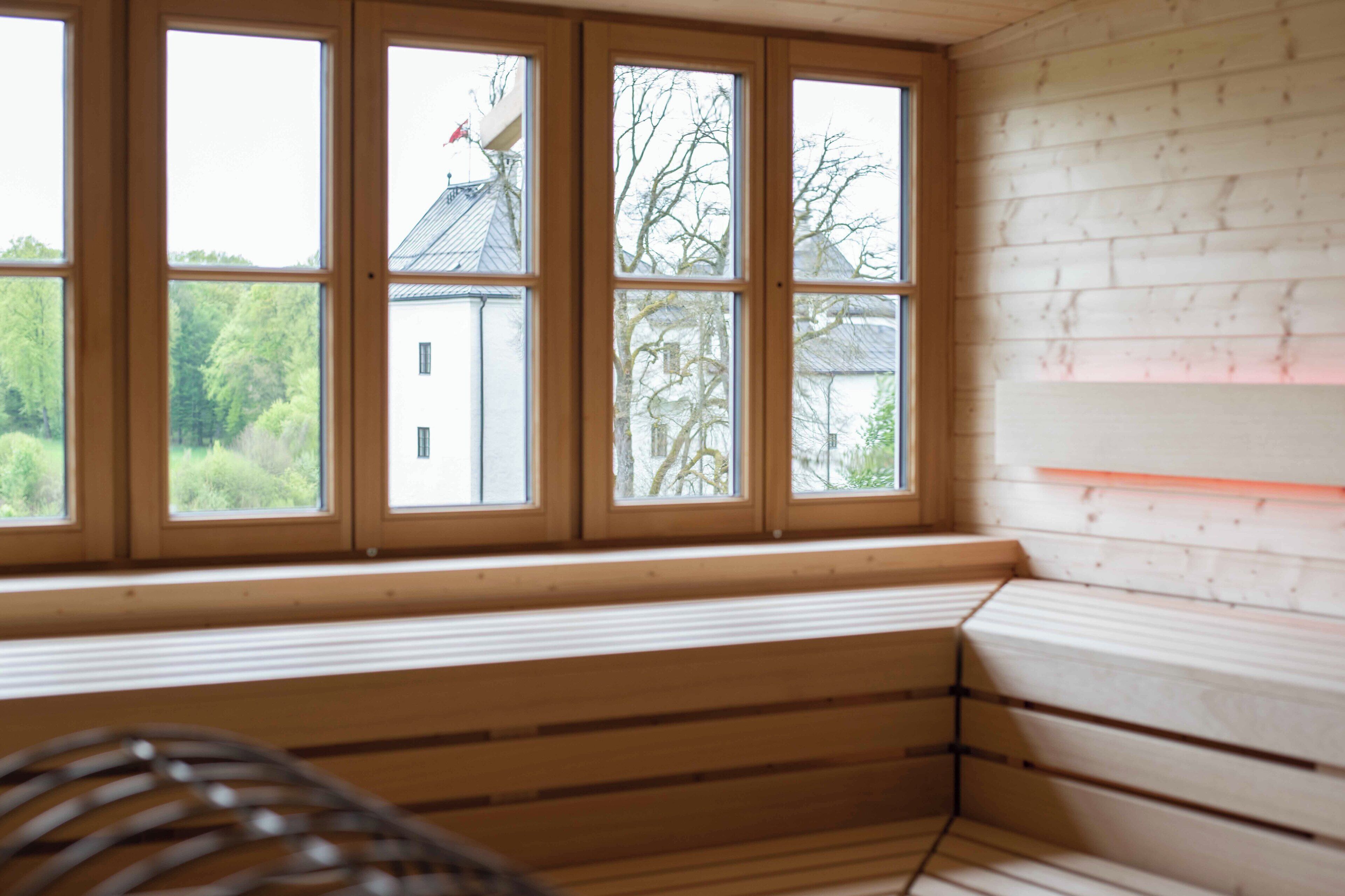 Een kamer met een houten bank bij een raam met veel ruitjes, een witte muur en een zichtbare boom buiten.