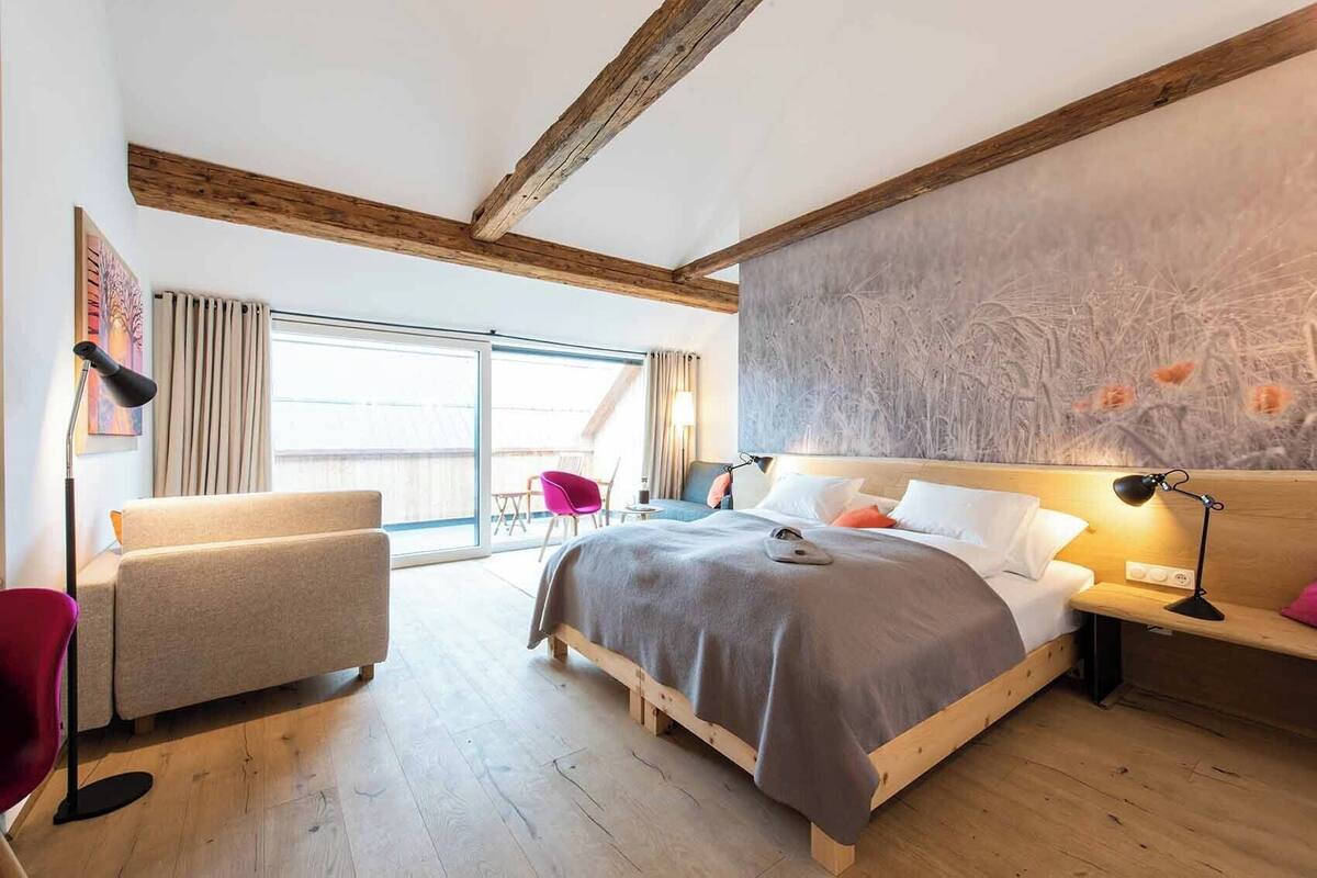 Ein modernes Schlafzimmer mit einem großen Bett, Holzbalken, einer Schiebeglastür zum Balkon, einem roten Sessel und einer Schreibtischlampe.