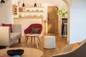 Un salon moderne avec un canapé, un fauteuil, une table ronde et un pouf. On aperçoit, au fond, un comptoir en bois doté d’étagères et d’une porte.