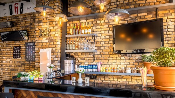 Um bar com parede de tijolos, prateleiras cheias de garrafas, uma televisão e um vaso de planta.