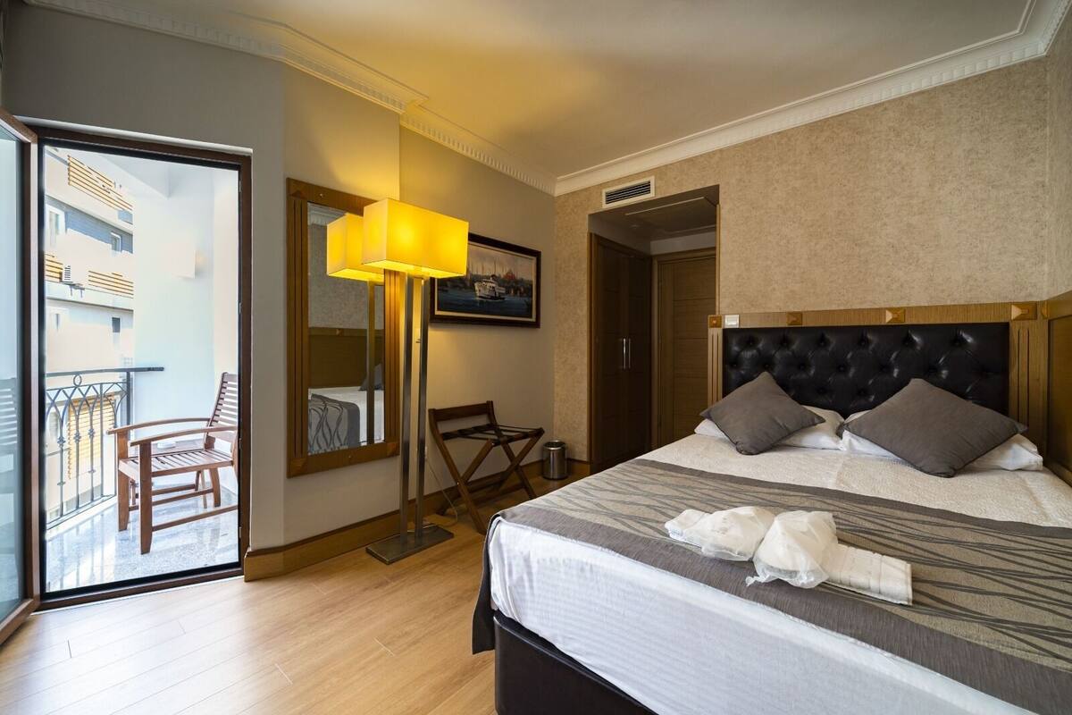 A hotel room with a bed, a chair, a desk, a lamp, a mirror, and a view of the outside.