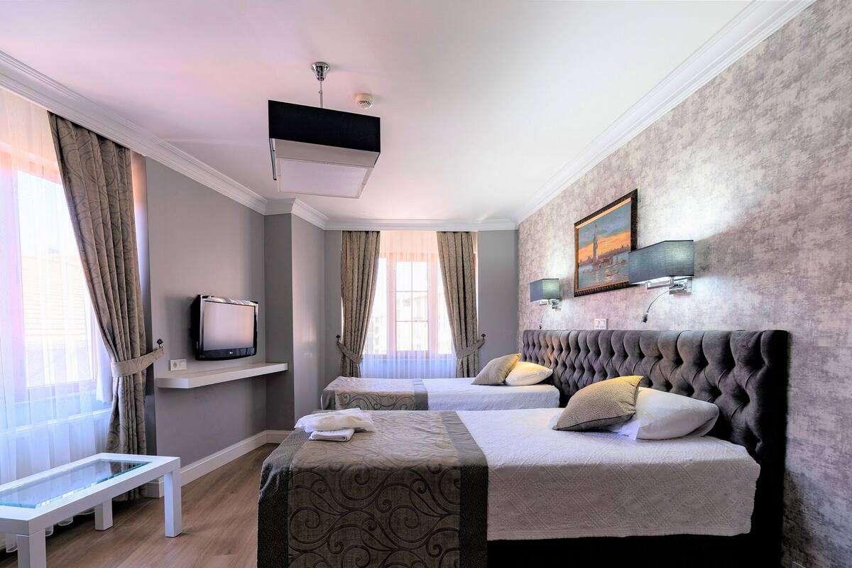 A hotel room with two beds, a television on a shelf, a painting on the wall, and a bench by the window.