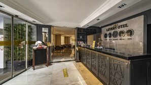 Um saguão de hotel com balcão de recepção, divisória de vidro e parede com relógios indicando diferentes cidades.